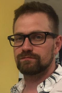 A man with glasses makes a serious expression at the camera