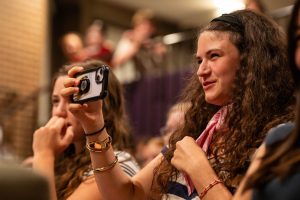 A person smiles as they film with a phone.