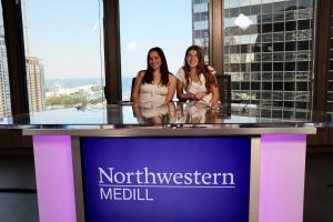 Two people sit behind a purple desk that says "Northwestern Medill" on the front. There are buildings and a river behind them.