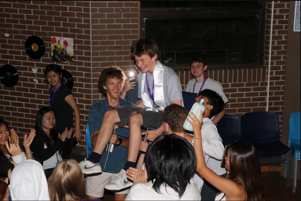 Two people in a crowd lift a chair with another person on it.