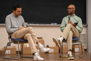 Two people sit on a stage with a blackboard and cardboard boxes in the background.