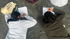 Two people lie on the ground and write on pieces of paper.